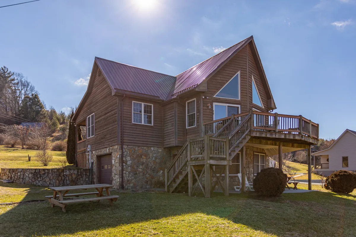 A spacious wooden deck extends from the house, featuring large windows and a stone foundation. The setting is configured for gatherings and outdoor meals, with views that suggest a seamless transition from indoor comfort to the wild mountain environment.
