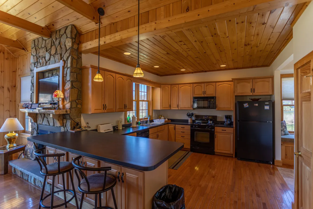 A warmly lit kitchen with a central island, stone fireplace in the background, ample cabinetry, and modern appliances. The space is open and inviting, ideal for gatherings and culinary activities. The kitchen island suggests both workspace and social hub, while the fireplace adds a cozy, communal vibe.