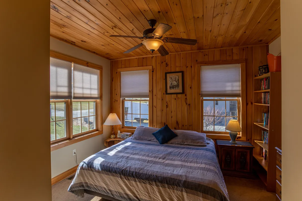 A serene bedroom highlighted by a comfortable bed with a blue accent pillow, wooden ceiling, ample windows letting in sunlight, a nightstand with a lamp, and a bookshelf. The scene is bright, restful, and thoughtfully designed.