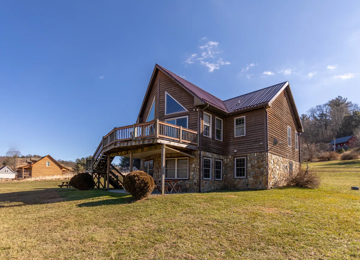 The image showcases the River House's inviting wooden deck overlooking the lush riverside, set against a charming stone exterior with large windows and a modern metal roof. The deck is perfectly suited for gatherings, offering panoramic views ideal for sunset watching, morning coffee, and evening barbecues with friends and family. The overall atmosphere is cozy and welcoming, framed by natural beauty and the promise of memorable outdoor moments.