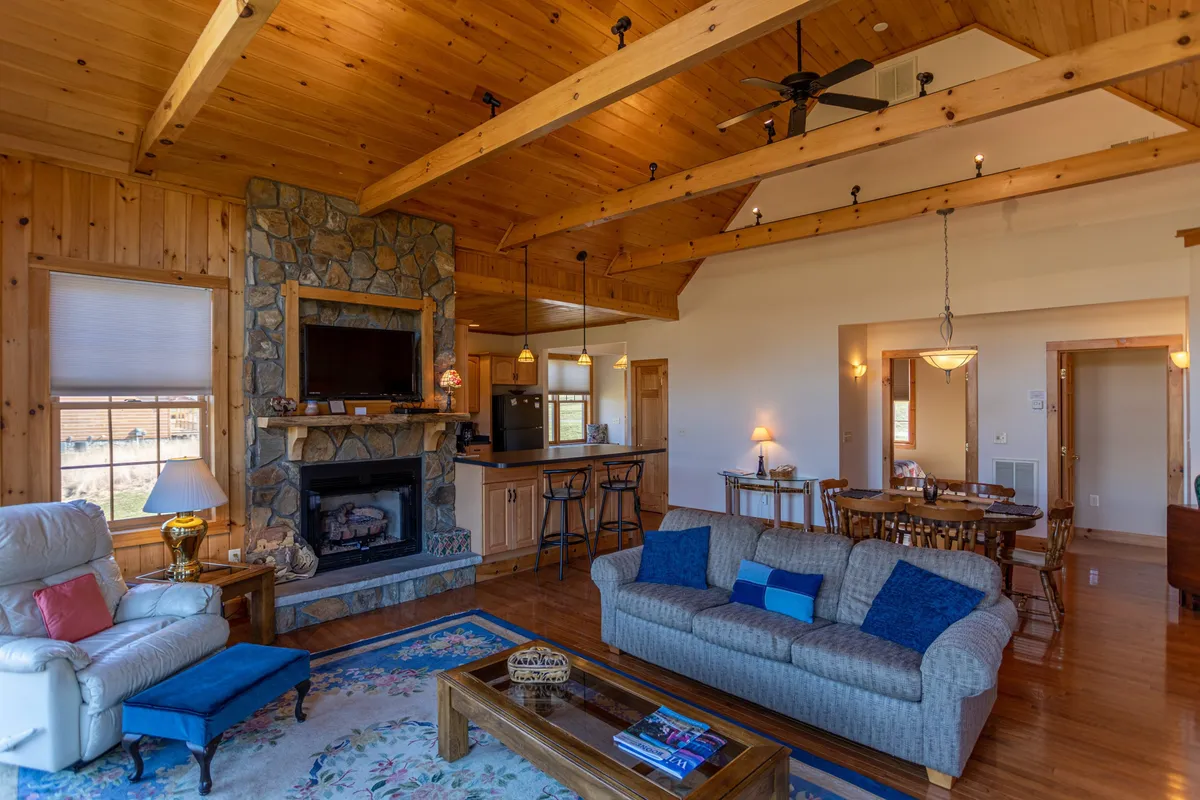 This image features a warm and inviting living room with a prominent stone fireplace, a cozy sofa accented with colorful cushions, a rustic dining table, and exposed wooden beams. The space exudes comfort and is clearly designed for gatherings and relaxation, with a welcoming atmosphere ideal for winter nights or family get-togethers.