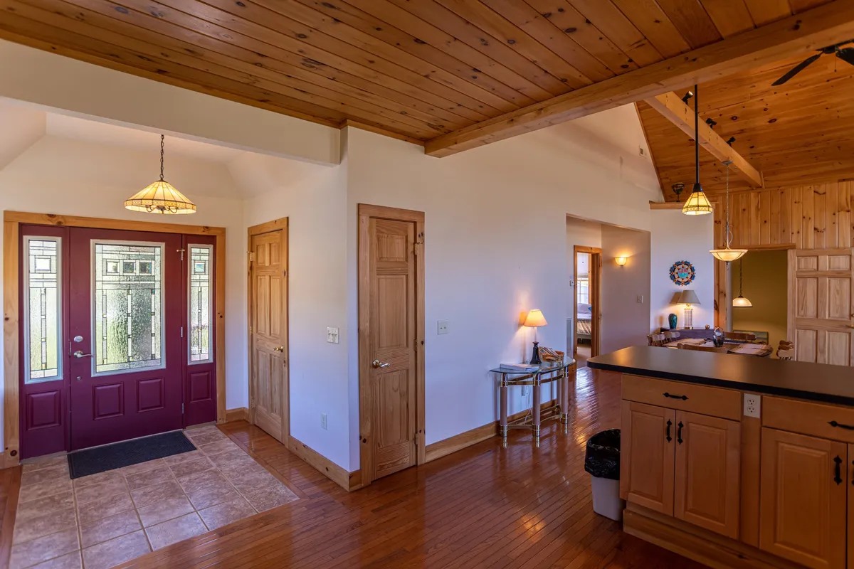 The image captures the entryway area, featuring a welcoming front door, a kitchen island, attractive wooden ceiling, and modern lighting fixtures. The space feels both practical and stylish, with clear sight lines that hint at a well-organized, functional entrance ideal for shedding boots or hanging dog leashes.
