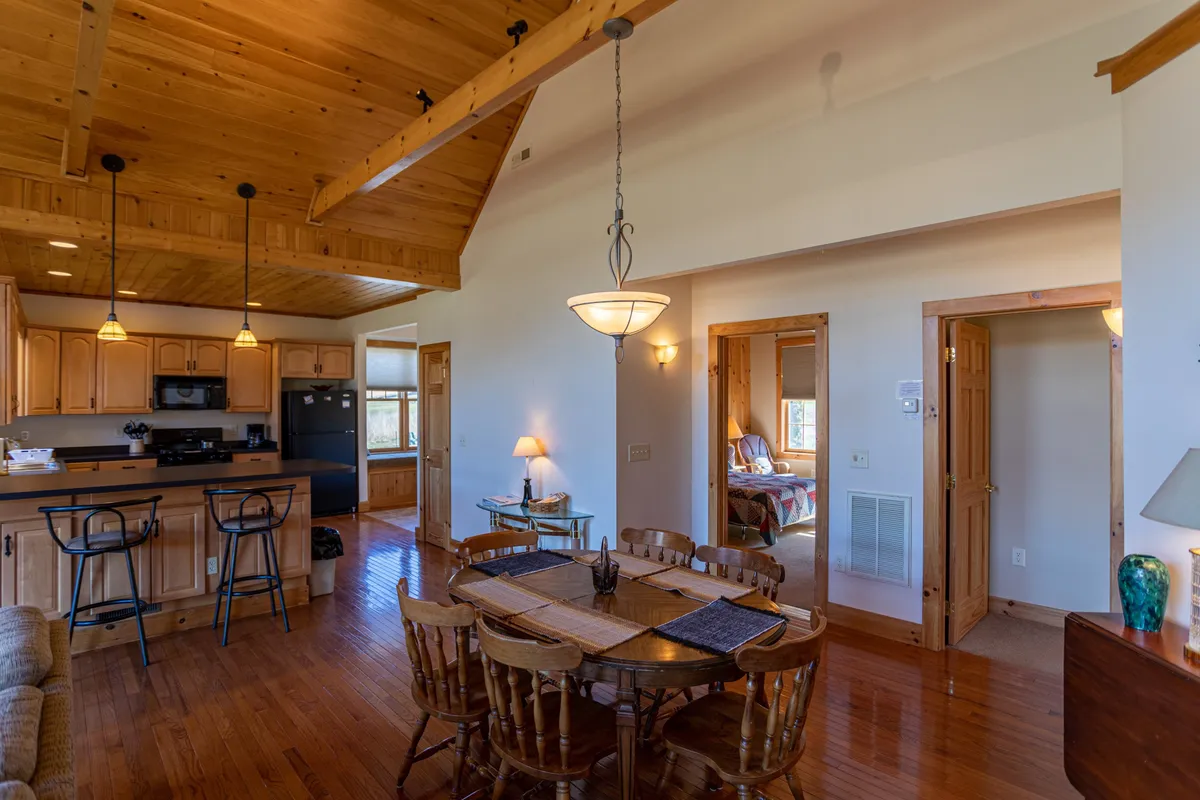 A cohesive open-plan interior featuring a wooden dining table adjacent to a kitchen bar and a cozy seating area, with large windows bringing in natural light and views of the outdoors. The arrangement highlights the flow between dining, kitchen, and living spaces.