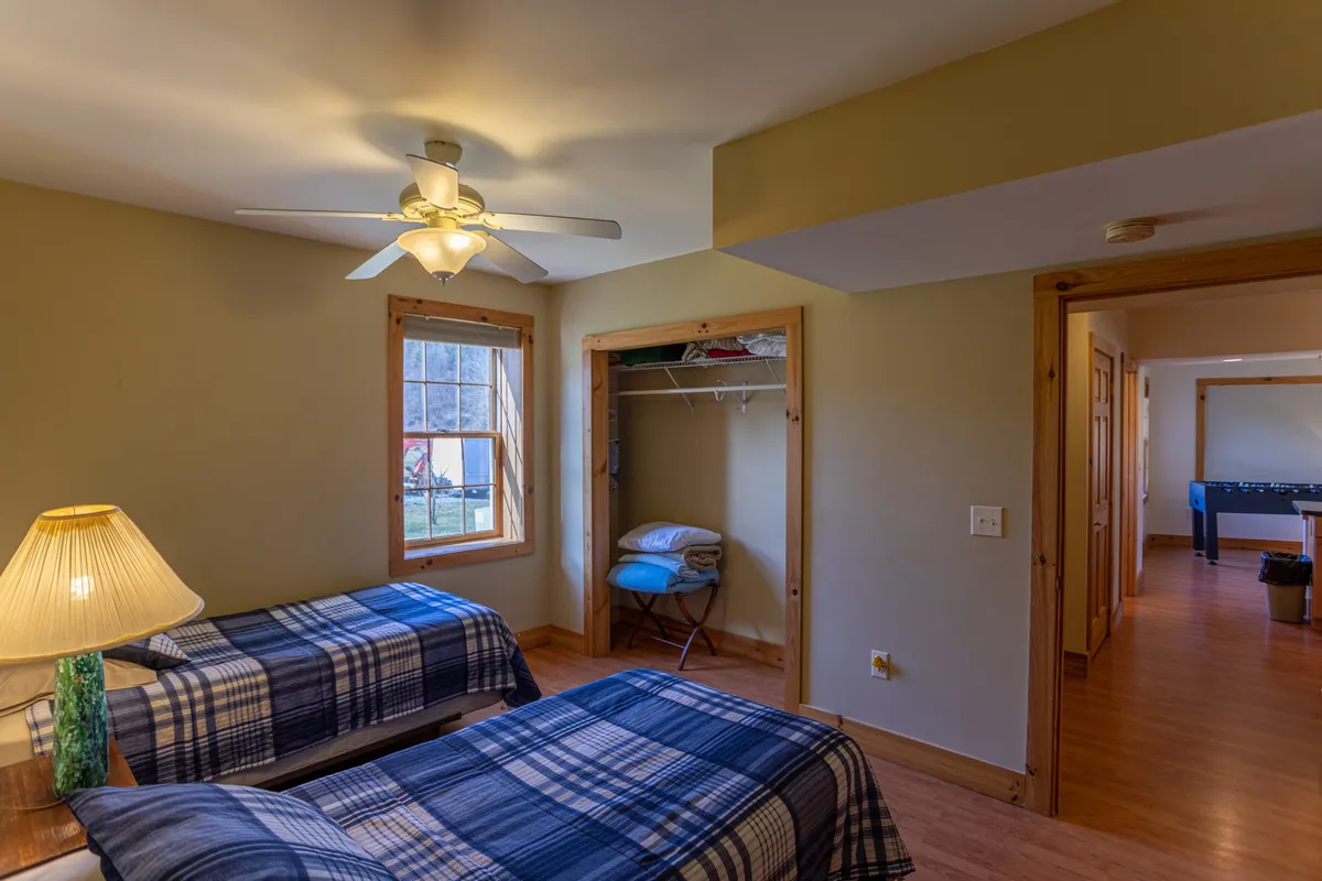 A spacious bedroom with two twin beds featuring plaid bedding, a ceiling fan overhead, a window allowing in natural light, and a bedside lamp. The arrangement is practical for siblings or a group of friends, with a sense of warmth and togetherness.