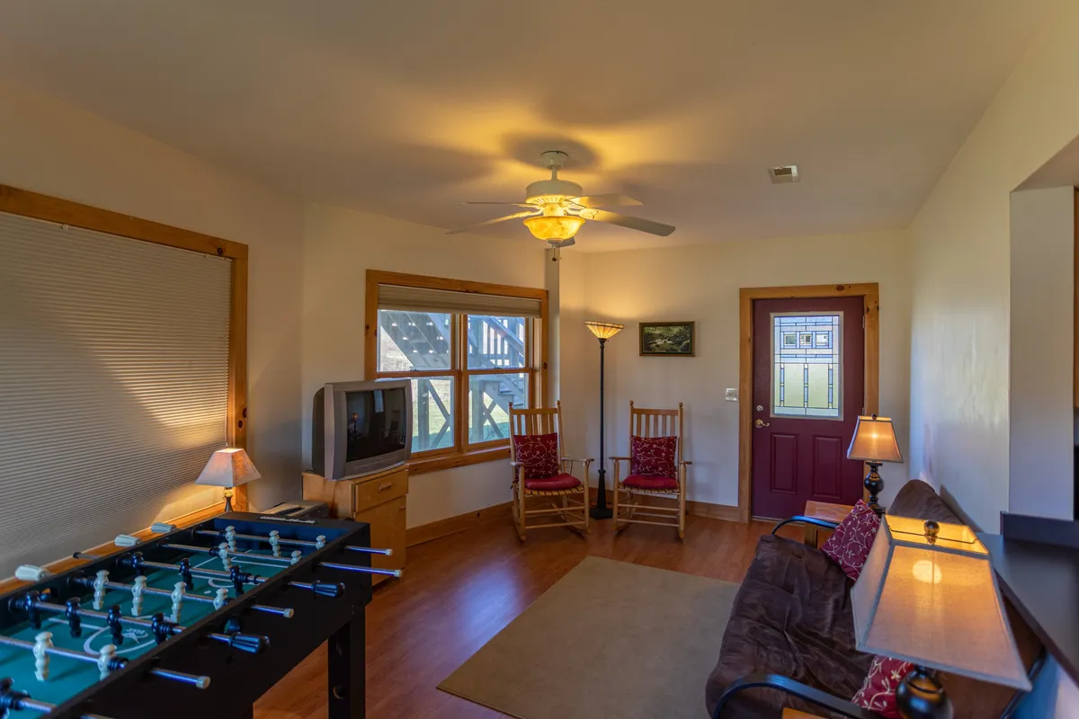 A lower-level lounge area equipped with a foosball table, a TV for movie nights, comfortable rocking chairs, and an entrance door hinting at easy access to the outdoors. The casual setting invites friendly competition, socializing, and relaxation for all ages.