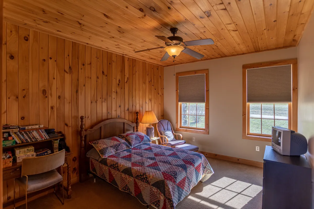 A welcoming bedroom featuring a cozy bed with a patterned quilt, rich wooden paneling, a plush reading chair, a warm lamp, and a bookshelf filled with books. The overall atmosphere is comfortable and inviting, ideal for relaxation and restful nights.