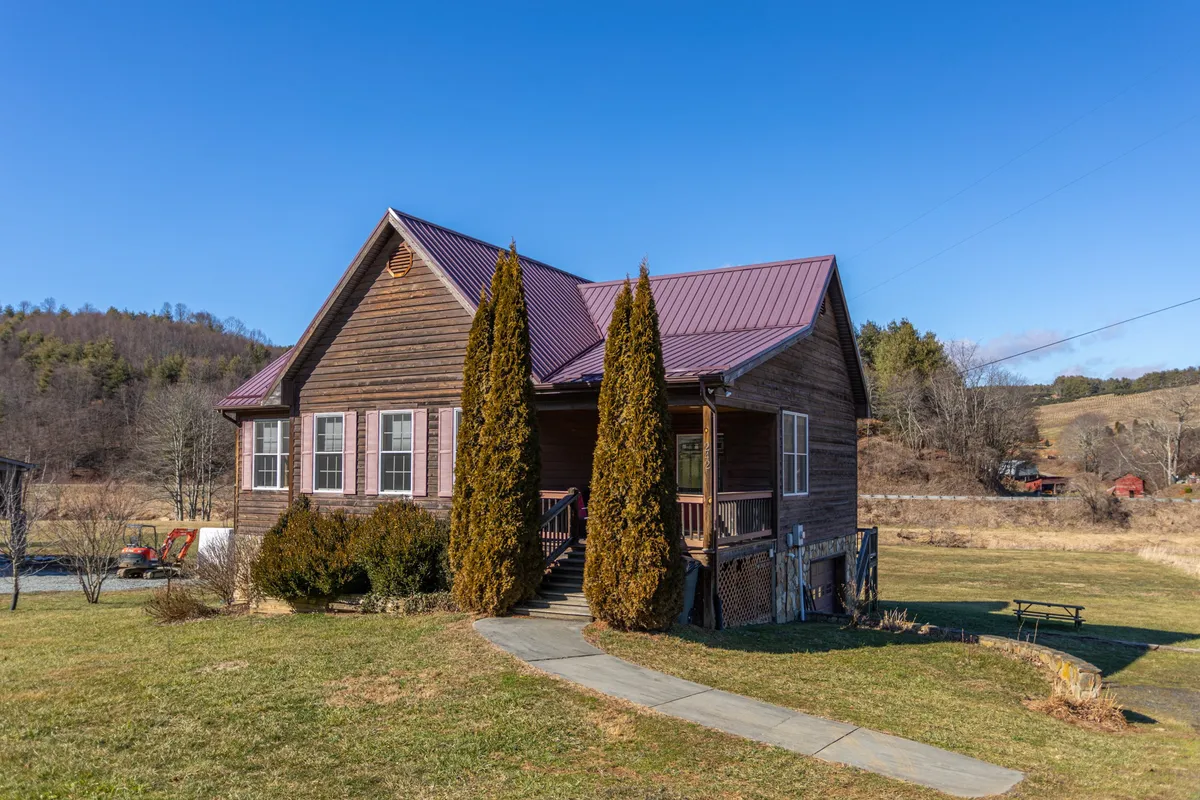 A welcoming wooden house framed by tall cypress trees, with a metal roof and inviting front steps. The large, flat yard in the foreground is ideal for outdoor activities, while people are seen relaxing on the porch and picnicking on the lawn, all set against a backdrop of peaceful mountain scenery.