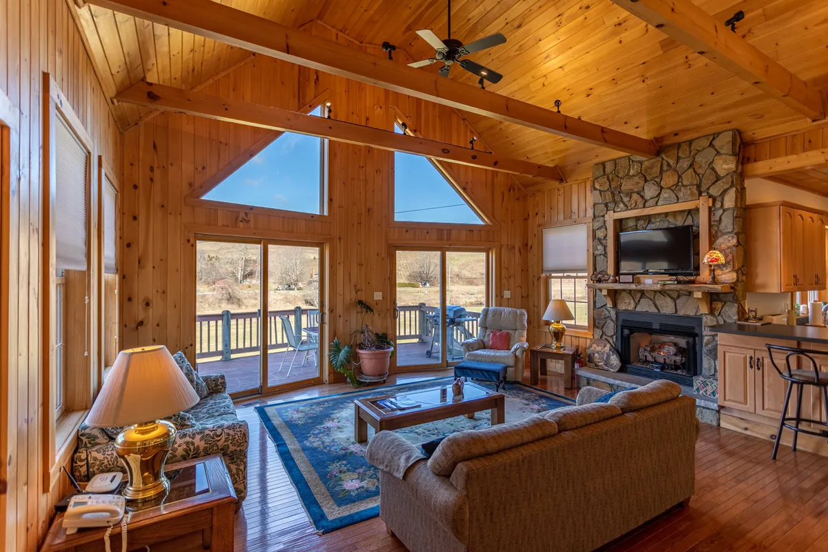 A living room defined by comfortable seating, a prominent stone fireplace, wooden beams overhead, and large windows offering stunning scenic vistas. The atmosphere is both welcoming and intimate, with visual cues for relaxation, conversation, and entertainment.