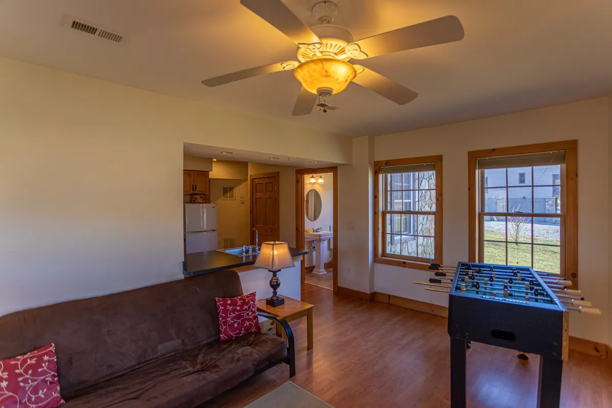 An inviting sitting area with a sofa decorated with colorful pillows, a foosball table in the foreground, a ceiling fan with soft lighting, and a window offering natural light and outdoor views. The layout is open and versatile, ideal for socializing, playing games, or enjoying quiet time.